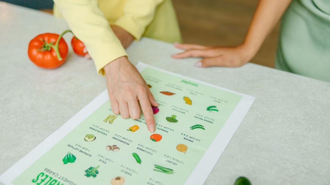 Close-up of hands pointing to a vegetable nutrition chart with fresh tomatoes on the table.